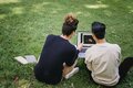 Zwei junge Menschen, die an einem Laptop sitzen, der vor ihnen auf einer Wiese steht.