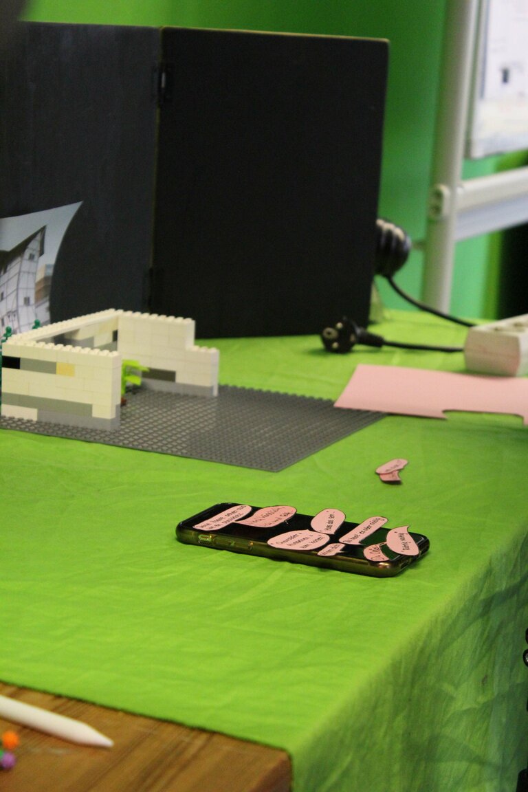 Ein Tisch, auf dem ein Smartphone mit Sprechblasen liegt. Zudem befinden sich eine Lego-Konstellation und ein Holzgestellt auf dem Tisch. 