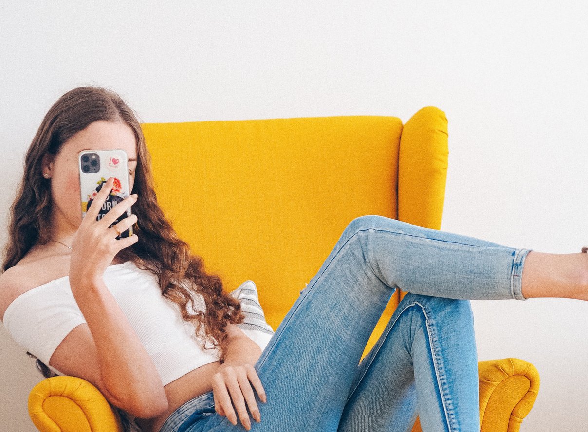 © Unsplash/Laura Chouette Eine Person, die auf einem Sessel sitzt und ein Smartphone vor ihr Gesicht hält.