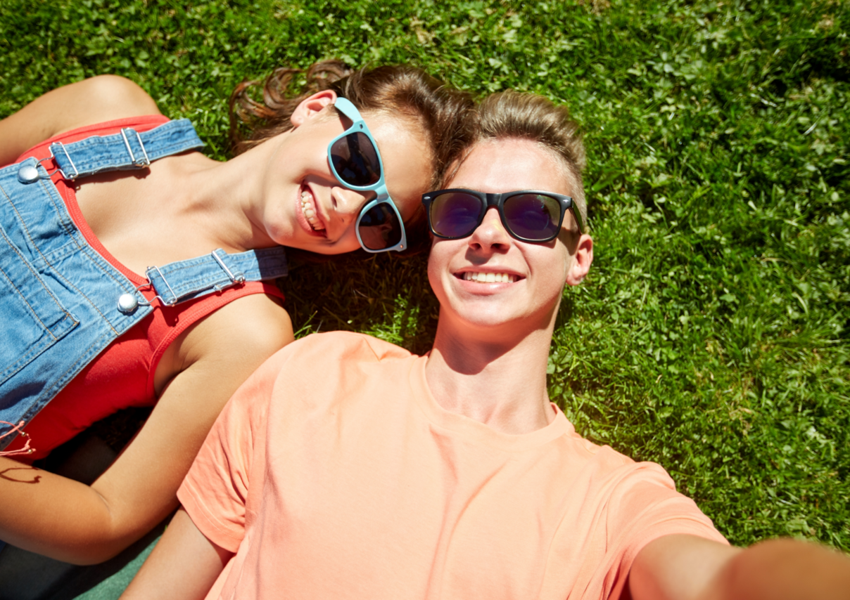 Zwei Teenager, die nebeneinander auf einer Wies liegen, Sonnenbrillen auf haben und in eine Kamera lachen.