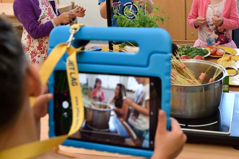 Ein Tablet, mit dem ein Kochtopf, welcher auf einem Herd steht, aufgenommen wird. Im Hintergrund sind Personen zu sehen, die Essen zubereiten.