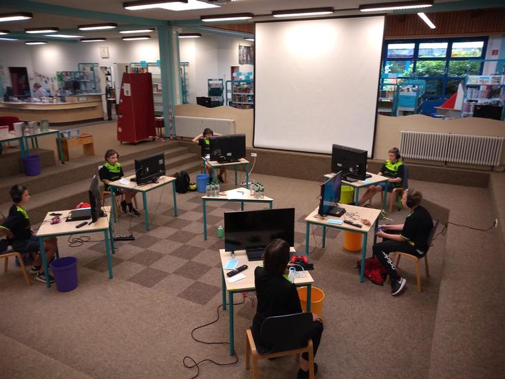 Kinder, die in einer Bibliothek an Tischen sitzen und einen PC vor sich stehen haben.