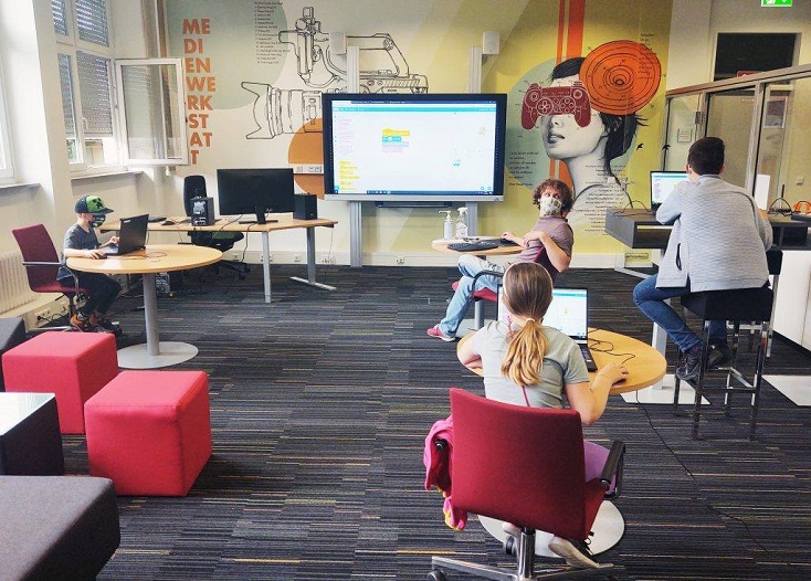 MoMo.jpg Computerspielschule Stuttgart: Die Lehrperson dreht sich um und blickt zu seinen drei Schüler:innen, welche jeweils an einem Tisch mit Laptop sitzen.