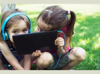 Zwei Kinder, die ein Tablet vor ihre Gesichter halten und auf einer Wiese sitzen. 