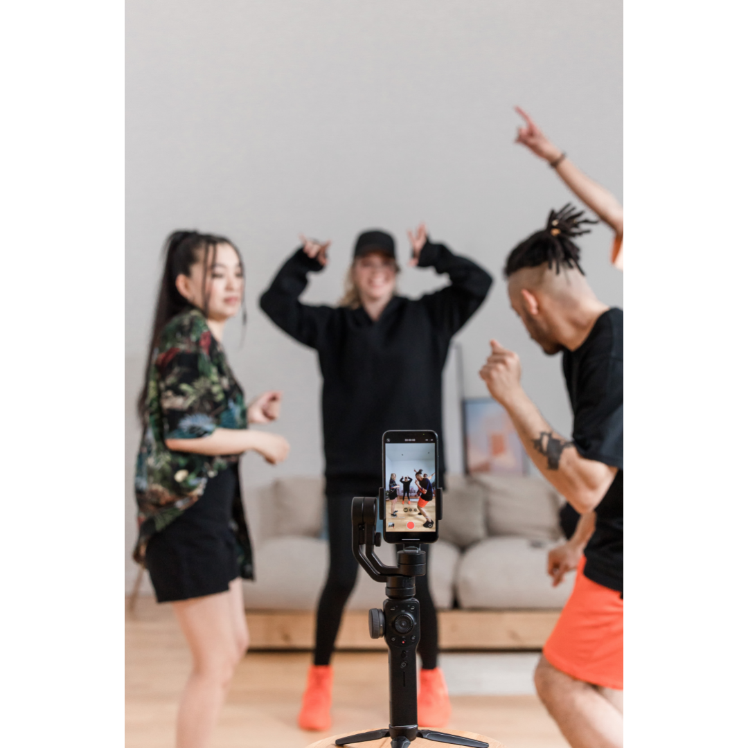 Drei Personen, die tanzen und dabei von einem Smartphone, welches auf einem Stativ befestigt ist, gefilmt werden. 